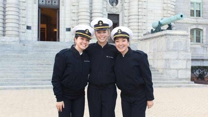 Three female cadets smile in front of a stone building.